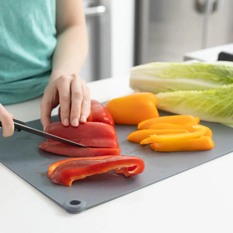 A person slices red and yellow bell peppers on the BPA-free, non-slip Karving King Supplement Cutting Board, with romaine lettuce nearby on a white kitchen counter.