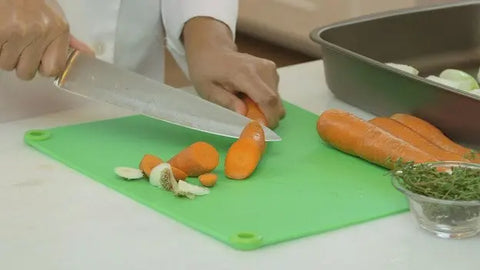 Using a Karving King Supplement Cutting Board, a person slices carrots with a large knife. Nearby are whole carrots, herbs in a small bowl, and a baking pan of vegetables. The board is green, non-slip, and BPA free.