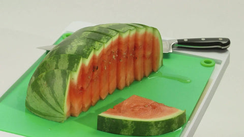 A sliced watermelon sits on the Karving King Supplement Cutting Board, with a kitchen knife in the fruit. One slice is separated in front, all set against a plain white background.