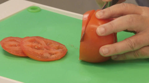 A hand is slicing a ripe tomato with a knife on the bright green, non-slip Karving King Supplement Cutting Board. Two tomato slices are beside the partially sliced tomato.