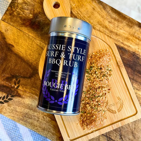 A tin from the Gustus Vitae Surf & Turf BBQ Seasonings Collection - 3 Pack sits on a wooden cutting board with a handle and engraved design, next to a pile of its herb-filled blend.