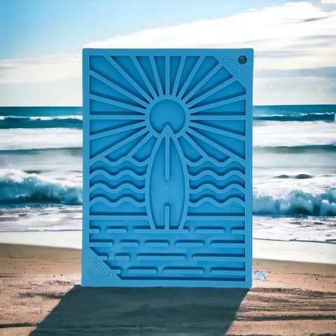 The SodaPup Surfs Up Design EMat Enrichment Lick Mat - Small, featuring sun, wave, and surfboard motifs, stands upright on a sandy beach under cloudy skies—ideal for canine enrichment and promoting foraging by the ocean.