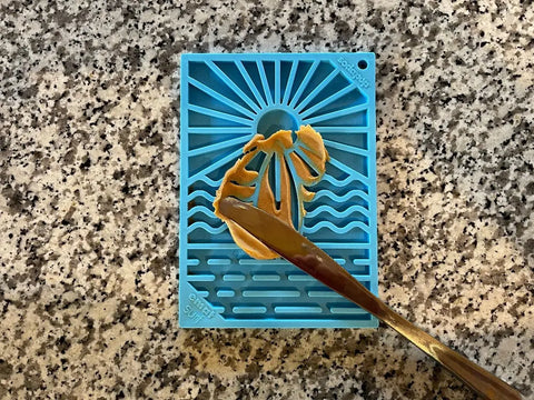 The SodaPup Surfs Up Design EMat Enrichment Lick Mat - Small features a sun and waves pattern, shown with peanut butter spread on it, ideal for canine enrichment and encouraging natural foraging instincts.