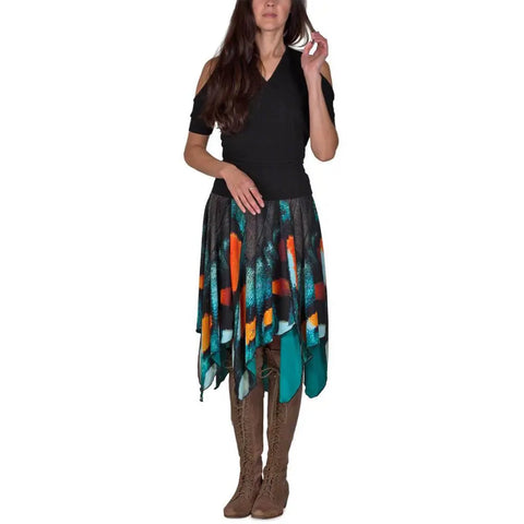 A woman stands against a white background wearing the Spiritex | Sustainable Apparel Mt. Pisgah Butterfly Skirt, featuring orange, teal, and black patterns, paired with a black cold-shoulder top and knee-high brown lace-up boots.