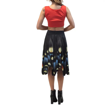 A woman stands facing away, wearing a red sleeveless crop top, black ankle boots, and the Spiritex Midnight Butterfly Skirt featuring a colorful abstract hem pattern.