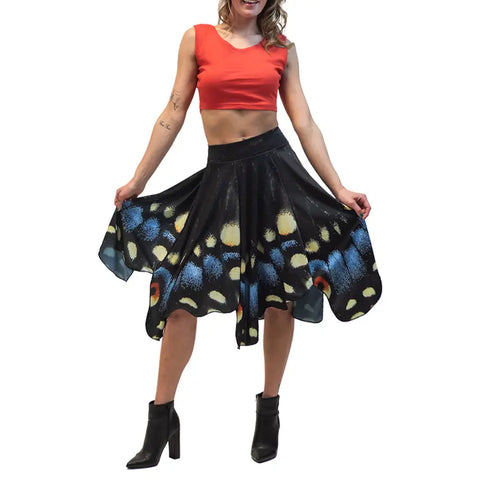 A woman wears a sleeveless orange crop top, black ankle boots, and the Midnight Butterfly Skirt by Spiritex | Sustainable Apparel, featuring hand-cut blue, yellow, and orange wing patterns.