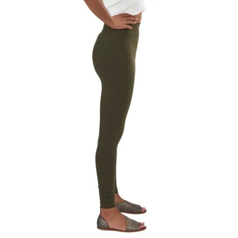 A person in profile stands against a plain white background, wearing Spiritex | Sustainable Apparel Yoga Leggings in olive green, paired with a white top and metallic-strapped, open-toed flat sandals.