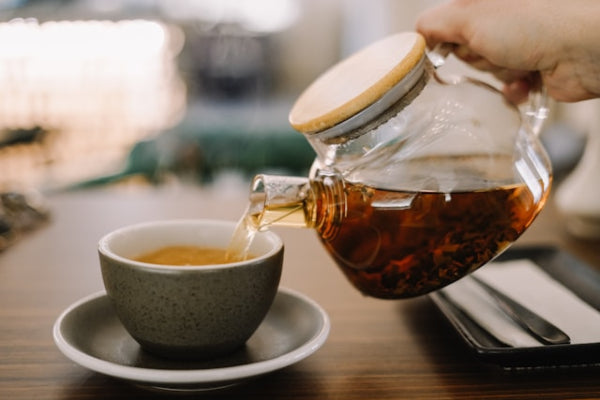 Tea being poured from a glass teapot into a ceramic cup on a wooden table.