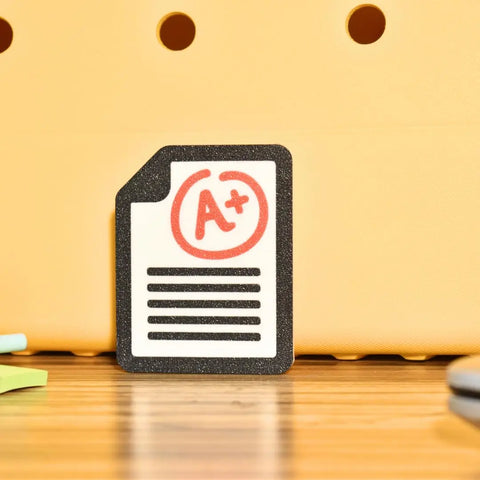 The FRESHe Baglets Teacher Charm by Chatelet Manufacturing, a decorative red A+ grade bag charm for tote bags like Bogg Bags and Simply Southern, is shown on a wooden surface with a yellow backdrop featuring circular holes.