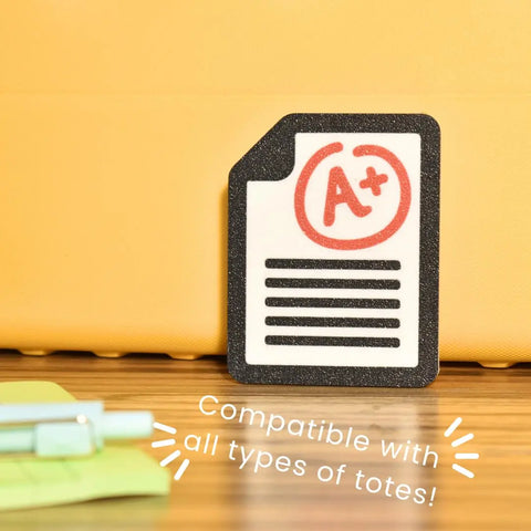 The FRESHe Baglets Teacher Charm by Chatelet Manufacturing features an A+ paper sticker in front of a yellow tote—perfect for teacher bags. Decorative and compatible with Bogg Bags, Simply Southern, and other totes! Pen and file folder also pictured.