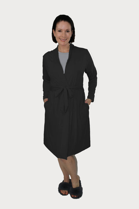 A woman smiles in the Goodnighties Banded Neck Robe—an American-made, black knee-length robe with a waist tie and pockets—styled over a gray top and paired with dark slippers, posing against a plain white background.