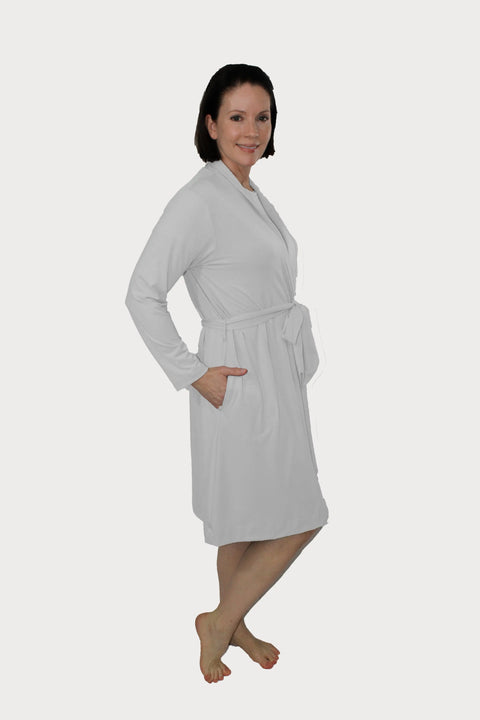 A smiling woman with short dark hair poses barefoot in the Goodnighties Banded Neck Robe, a knee-length, light gray American-made robe with long sleeves and a waist tie.