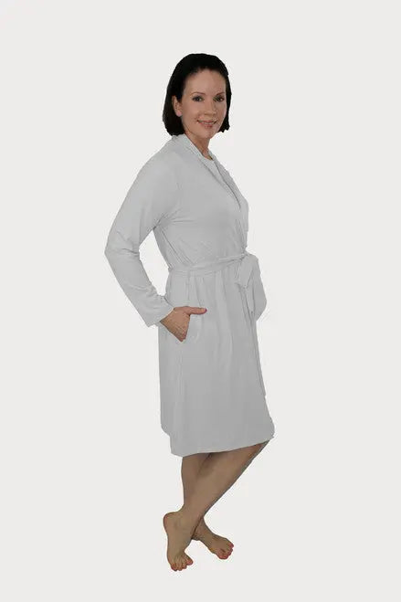 A woman with short dark hair smiles while wearing the Goodnighties Banded Neck Robe, an American-made light gray robe with a belt and pockets. The knee-length robe is shown as she stands barefoot against a plain white background.