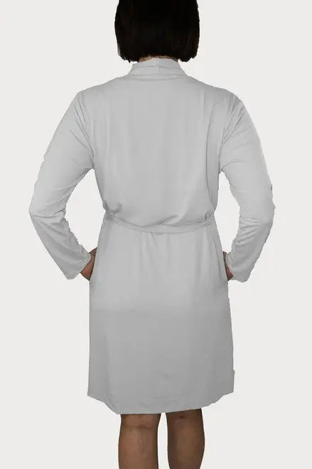 A person with short dark hair is seen from the back wearing the Goodnighties Banded Neck Robe, a knee-length, long-sleeve American-made robe tied at the waist. The background is plain and light-colored.