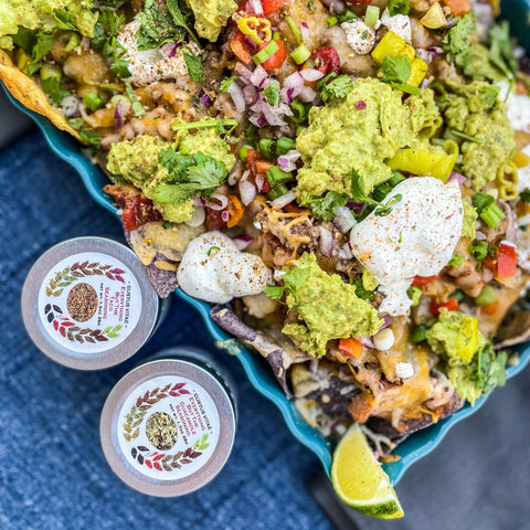 A vibrant plate of loaded nachos with guacamole, sour cream, onions, cilantro, and peppers is served with lime wedges. Beside it are two small containers from the Gustus Vitae Tex-Mex Flavors Seasoning Collection on a blue surface.