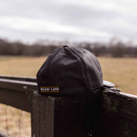 A black Gate Performance Stretch Fit Hat by Wilson Lane Apparel rests on a dark wooden fence post, with an open field and cloudy sky in the background.