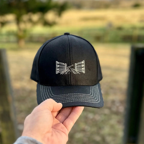 A hand holds The Gate Performance Stretch Fit Hat by Wilson Lane Apparel—a black cap featuring a white embroidered two gates handshake design. The blurred rural background shows lush green grass and trees.