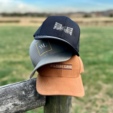 Three Wilson Lane Apparel The Gate Performance Stretch Fit Hats—black, gray, and tan with unique embroidered logos—are stacked on a wooden fence in a grassy field against a blurred landscape.