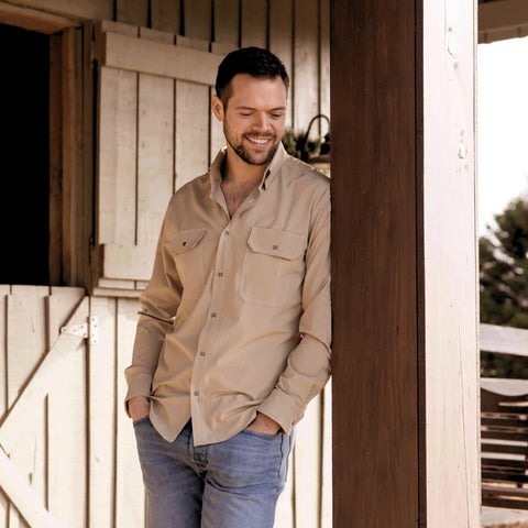A man with short dark hair and a beard, wearing the Nash Button Down Shirt - Desert by Wilson Lane Apparel and blue jeans, stands smiling and leaning against a wooden post in front of a barn.
