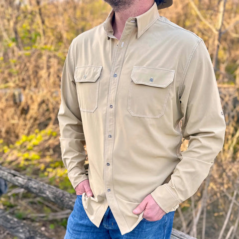 Wearing The Nash Button Down Shirt in Desert by Wilson Lane Apparel and blue jeans, a person stands outdoors near a wooden fence with hands in pockets, surrounded by trees and foliage in soft natural light.