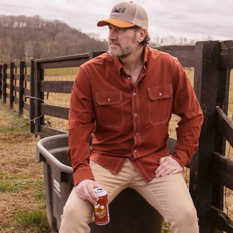 A man wearing the Wilson Lane Apparel Nash Button Down Shirt in Brick and tan pants sits on a large plastic bin by a wooden fence, holding a drink can, with hills and rural scenery in the background.
