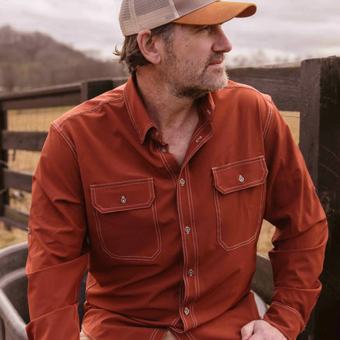 A man in Wilson Lane Apparel’s The Nash Button Down Shirt - Brick and a tan cap sits outdoors by a wooden fence, looking to the side with a relaxed expression.