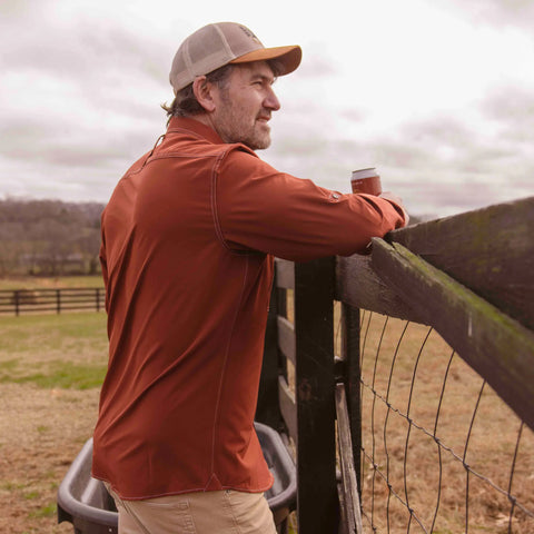 A man in a brown jacket and cap, wearing the Wilson Lane Apparel Nash Button Down Shirt in Brick, leans on a wooden fence with a can, gazing over fields beneath cloudy skies.
