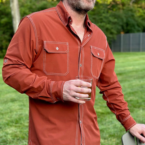 A man outdoors on grass wears the Wilson Lane Apparel Nash Button Down Shirt in Brick, holding a glass of brown liquid and resting his arm on a chair. Trees and a fence can be seen in the background.