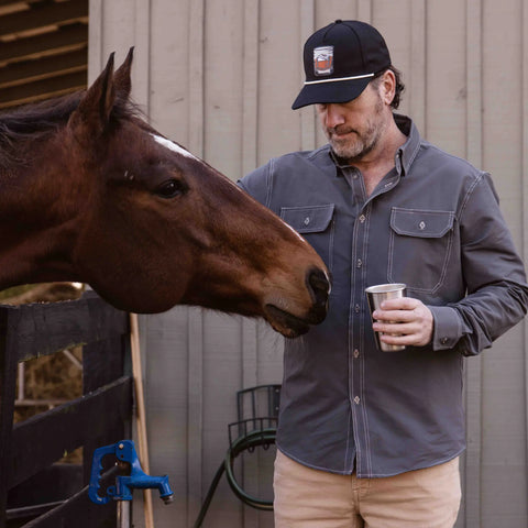 Wearing the Wilson Button Down Shirt - Ash by Wilson Lane Apparel and a black cap, a man stands by a brown horse, gently touching its muzzle while holding a metal cup near a wooden building and fence.