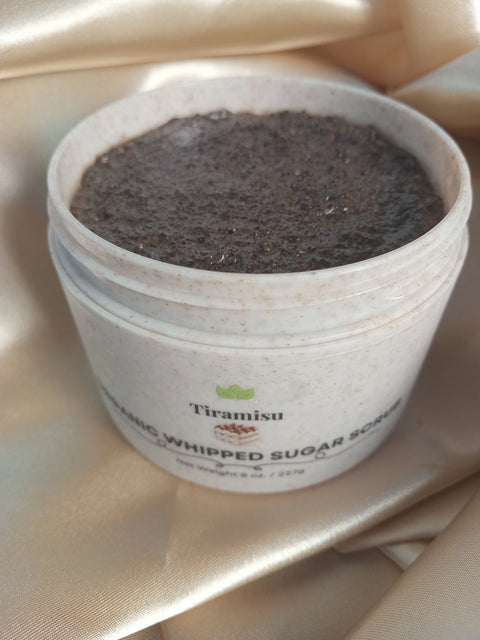 An open jar of Forev'Herbs Tiramisu Sugar Scrub rests on silky beige fabric, showcasing its textured brown sugar scrub. The white container with green and brown labels highlights this coffee body scrub's elegance and indulgence.