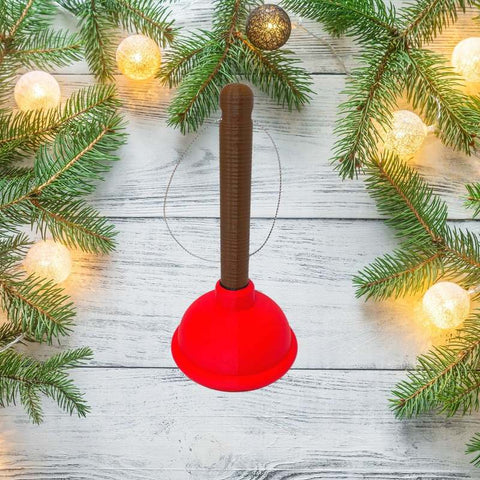 The Toilet Plunger Christmas Ornament by Chatelet Manufacturing, a funny holiday decor item made in the USA, is shown on a white wooden surface with pine branches and string lights for a festive touch.