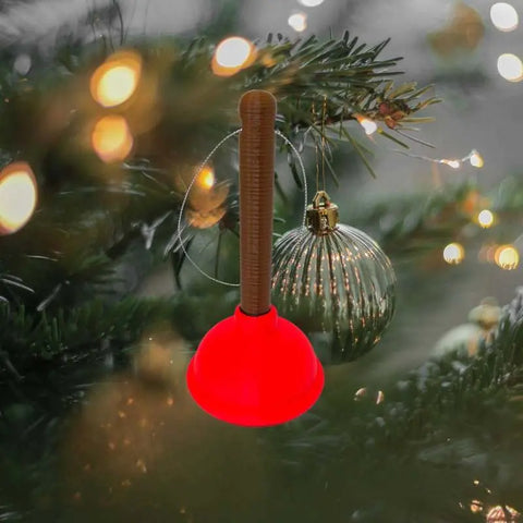 The Chatelet Manufacturing Toilet Plunger Christmas Ornament, a funny holiday decoration made in the USA, hangs on a tree next to a gold-striped bauble with warm lights glowing softly in the background.