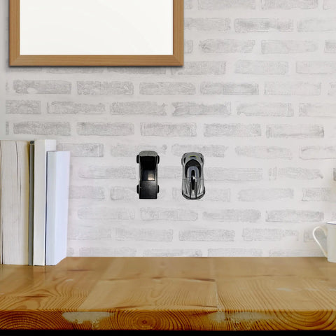 A wooden desk with books and a white mug sits before a white brick wall. Above, two black hooks hold a Chatelet Manufacturing Toy Car Wall Display Organizer, perfect for displaying Hot Wheels beneath a wooden-framed mirror.