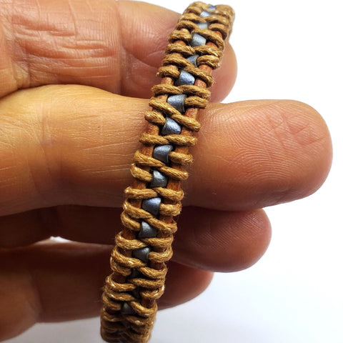A hand holds the Tree of Life Button Brown Gray Leather Wrap Seed Bead Bracelet by Alexa Martha Designs, featuring brown cord woven around metallic silver beads for a textured, sturdy style.