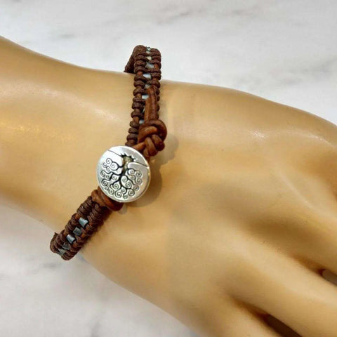 The Tree of Life Button Brown Gray Leather Wrap Seed Bead Bracelet by Alexa Martha Designs features a round silver tree of life charm on braided leather, shown on a mannequin hand against a light background.