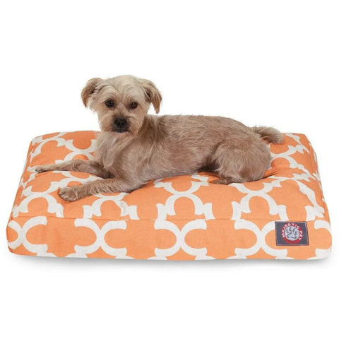A small, light brown dog lies on the Majestic Pet Trellis Rectangle Dog Bed, featuring an orange and white pattern with a logo patch. Its premium polyester fill offers comfort and the washable cover makes cleaning easy. The dog looks towards the camera.