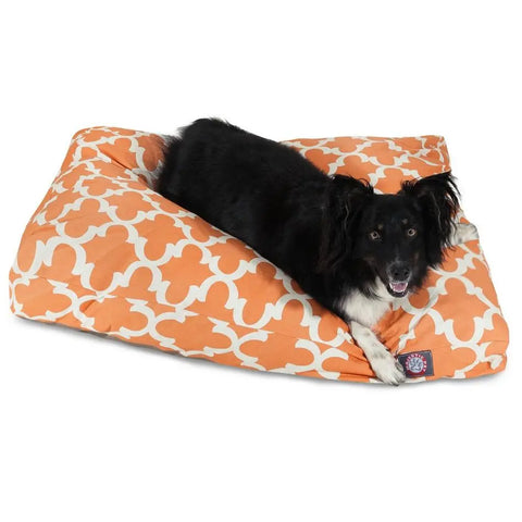 A black and white dog lies on a Majestic Pet Trellis Rectangle Dog Bed with premium polyester fill, featuring an orange and white pattern, looking towards the camera with its mouth slightly open.