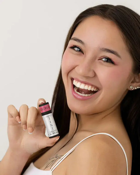 A smiling woman with long dark hair holds Cheryl Kelly & Co.’s Triple Tint Therapy—Lip, Cheek & Eye Color—in eco-friendly packaging near her face. She wears light makeup, pink blush and eyeshadow, subtle lip tint, and a white tank top.