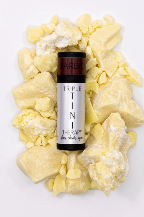 A silver tube of Cheryl Kelly & Co. Triple Tint Therapy - Lip, Cheek & Eye Color with a dark cap sits on scattered yellow cocoa butter, spotlighting its plant squalane lip tint formula against a white background.