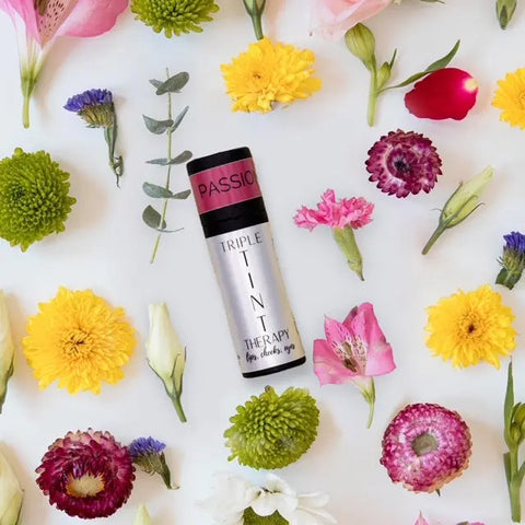 A tube of Cheryl Kelly & Co. Triple Tint Therapy – Lip, Cheek & Eye Color in “Passion” sits among vibrant blooms on a white surface, highlighting its plant squalane formula and eco-friendly packaging.