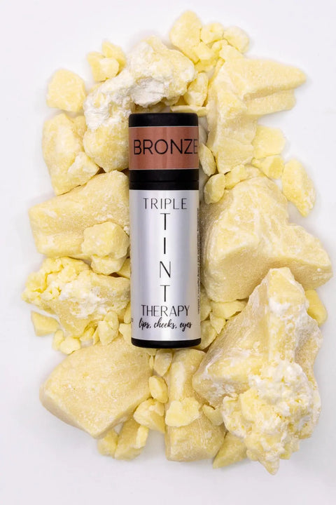 A tube of Cheryl Kelly & Co. Triple Tint Therapy - Lip, Cheek & Eye Color in bronze shade, enriched with plant squalane, sits on yellow shea butter chunks. This versatile tint is suitable for lips, cheeks, and eyes.