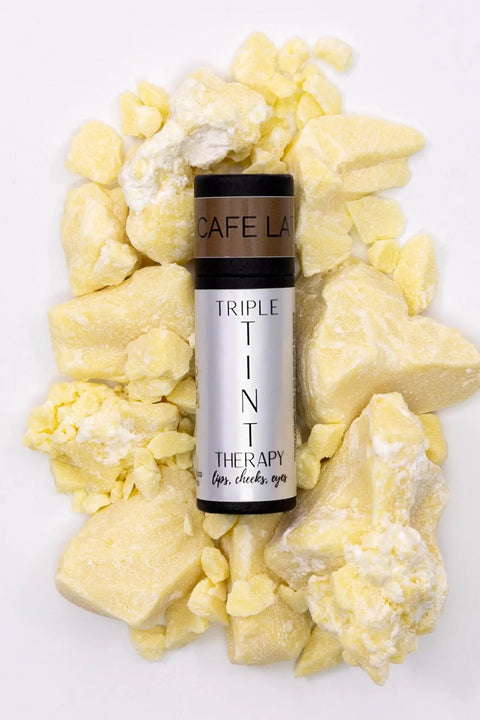 A silver tube of Cheryl Kelly & Co. Triple Tint Therapy - Lip, Cheek & Eye Color in Cafe Latte sits among chunks of yellowish, crumbly raw butter on a white background, highlighting its eco-friendly packaging.