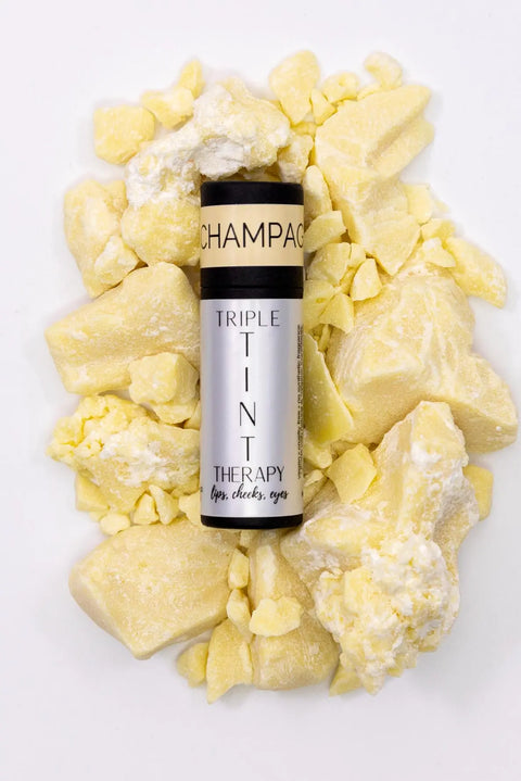 A silver tube labeled Triple Tint Therapy - Lip, Cheek & Eye Color by Cheryl Kelly & Co. rests on chunky yellow and white solids, highlighting its plant squalane formula. The cap is marked CHAMPAGNE.