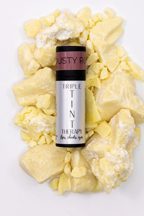 A tube of Cheryl Kelly & Co. Triple Tint Therapy - Lip, Cheek & Eye Color rests on chunky raw shea butter, highlighting its nourishing plant squalane formula and eco-friendly packaging against a white background.