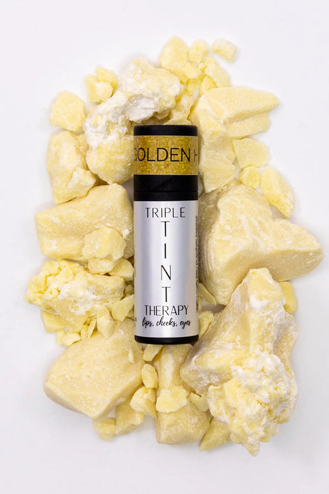 A tube labeled Triple Tint Therapy - Lip, Cheek & Eye Color by Cheryl Kelly & Co. rests on yellow shea butter pieces atop a white background. This versatile product comes in eco-friendly packaging.