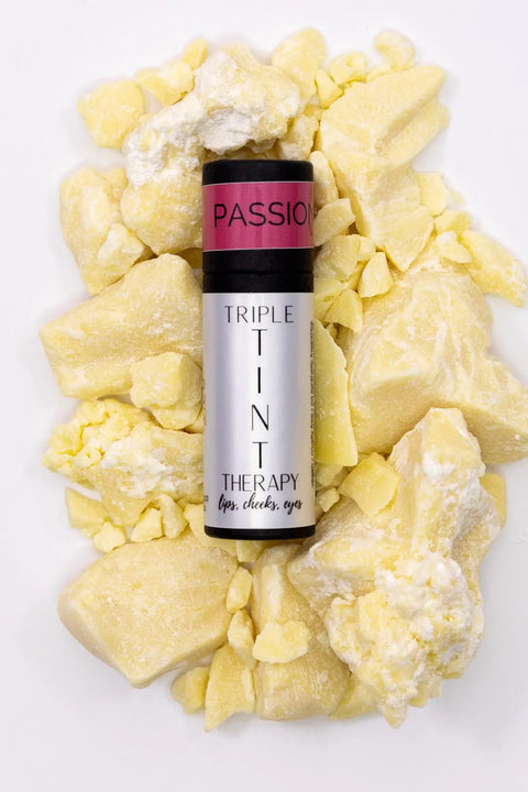 A silver tube of Cheryl Kelly & Co. Triple Tint Therapy - Lip, Cheek & Eye Color rests on scattered yellow cocoa butter chunks, highlighting plant squalane and eco-friendly packaging.