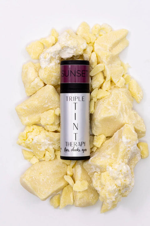A tube of Cheryl Kelly & Co. Triple Tint Therapy - Lip, Cheek & Eye Color in SUNSET rests on chunky yellow balm, set against a white background—highlighting its eco-friendly packaging.