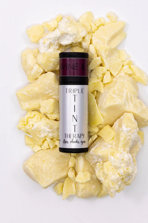 A tube of Cheryl Kelly & Co. Triple Tint Therapy - Lip, Cheek & Eye Color sits on chunky raw shea butter. Infused with plant squalane and in eco-friendly packaging, it stands out against a crisp white background.