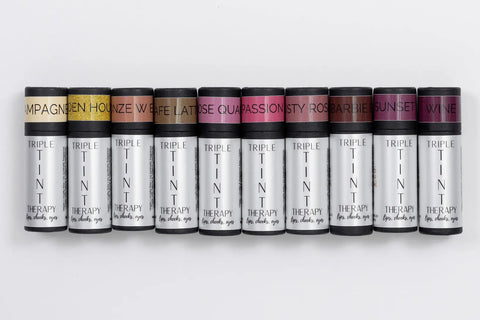 Ten tubes of Cheryl Kelly & Co. Triple Tint Therapy - Lip, Cheek & Eye Color, each with a distinct colored cap and flavor label like Champagne, Honey, and Wine, are arranged side by side in eco-friendly packaging with plant squalane.
