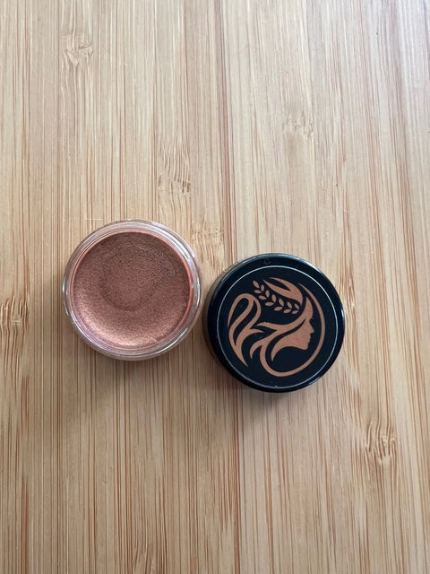 A small jar of Cheryl Kelly & Co. Triple Tint Therapy - Lip, Cheek & Eye Color in shimmery copper rests on a wooden surface, its black lid with a copper woman’s profile and wheat design placed beside it.